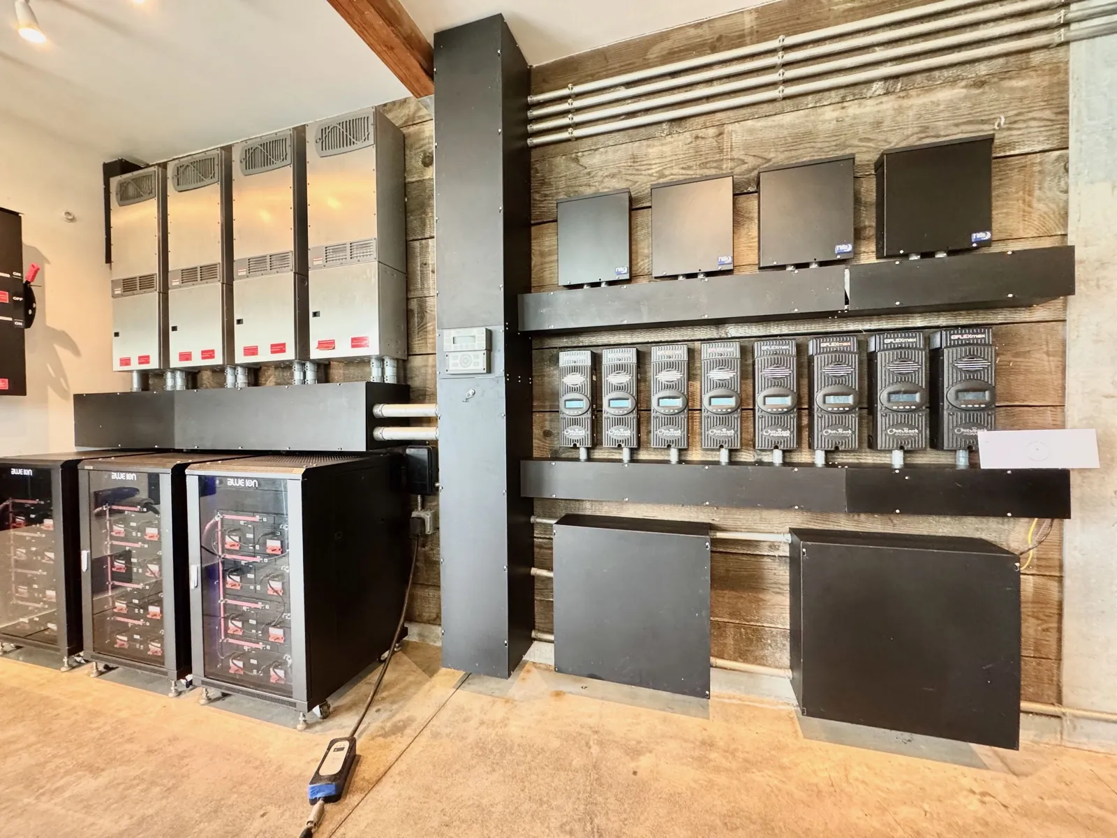 Close-up of the off-grid power wall: Blue Ion lithium battery banks below a row of OutBack FLEXmax charge controllers and inverters, mounted on reclaimed wood