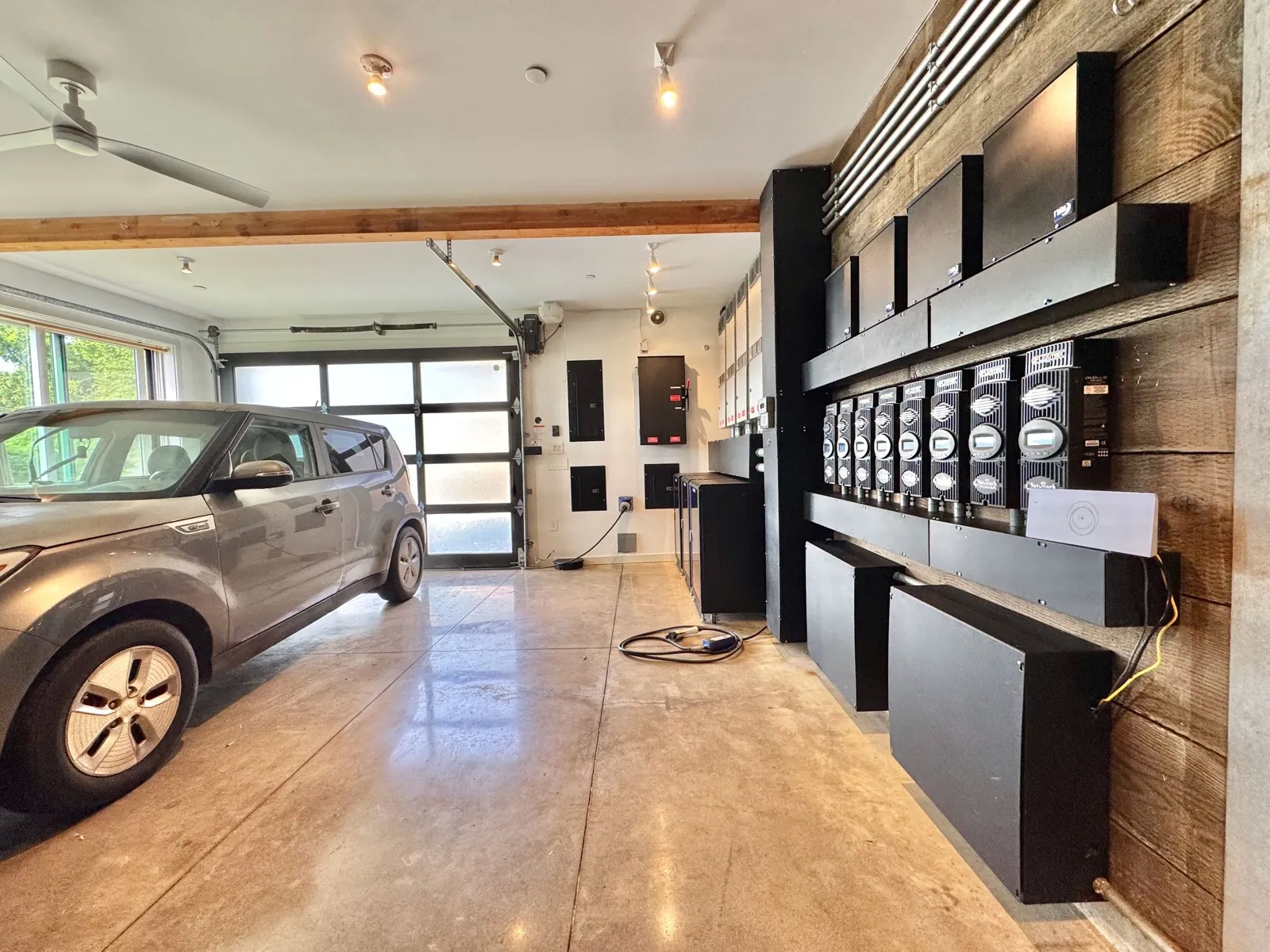 Garage interior: car parked beside the frosted-glass roll-up door, with the full off-grid power wall — OutBack inverters and charge controllers on reclaimed-wood cladding — along the right