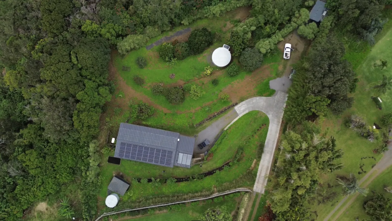 Top-down birdseye view of the property: standing-seam roof with solar array, water tank, geodesic dome, the curving driveway and surrounding food forest