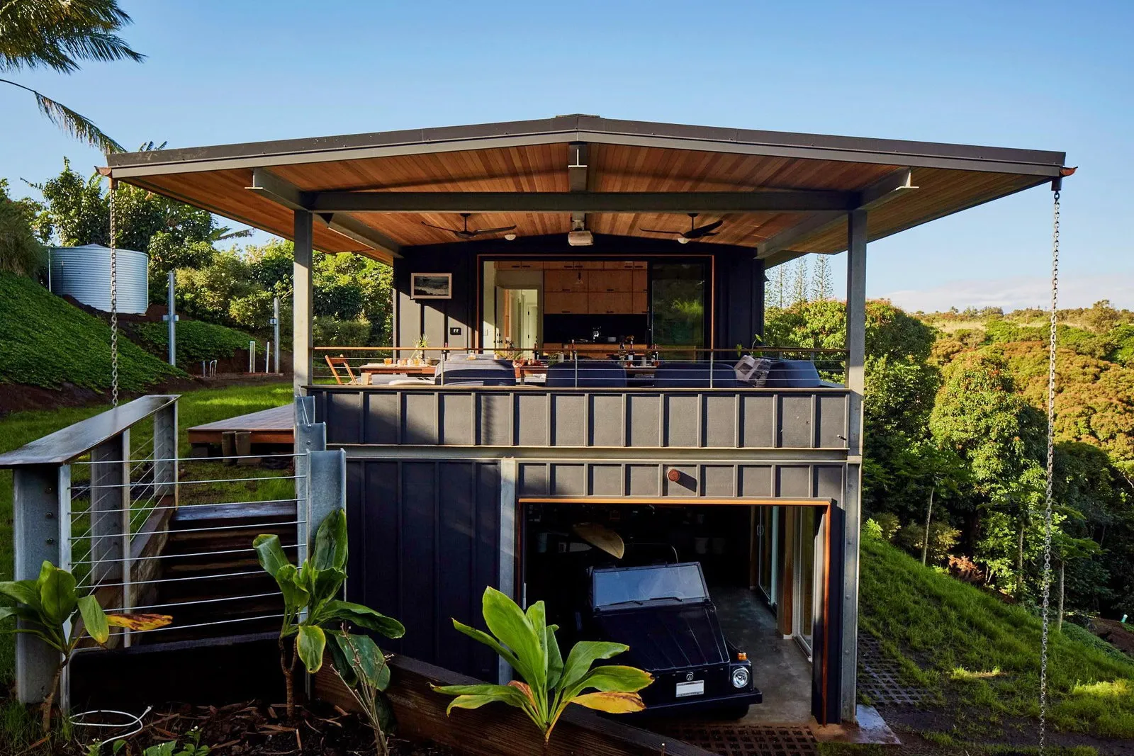 Back of the house at golden hour — wide cedar-soffit roof, upper lanai dining, lower garage with the VW Thing peeking out, water tank chains on the side